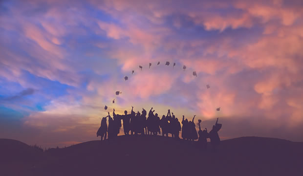 Students standing on hilltop in cap and gown throwing caps